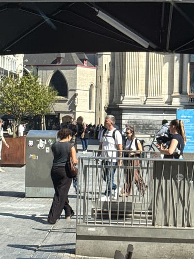 The image shows a street scene in Brussels, Belgium, with several people present. In the foreground, a person in a black shirt and black pants is lying on the ground with their legs extended. Behind them, a gray trash bin is visible, adorned with several stickers. To the left, a large, dark structure, possibly a stage or canopy, dominates the frame.
Further back, a group of people are standing or walking near a metal fence. Among them, a person with long blonde hair is wearing a black t-shirt and jeans, leaning forward. Another person in the background wears a white t-shirt. The architectural style of the buildings suggests an urban European setting, consistent with Brussels. The sunlight indicates it is daytime, likely with clear weather, casting shadows on the ground. The overall atmosphere appears to be relaxed, possibly a public gathering or event. No visible text is discernible in the image.