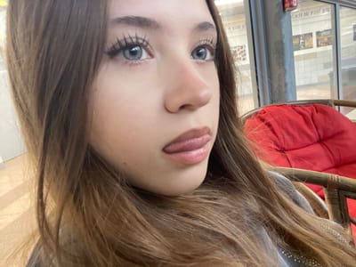 A close-up portrait image captures a young female subject positioned slightly off-center to the left. She has long, light brown hair, which partially obscures the left side of her face and extends over her shoulders. Her visible right eye has a light blue iris, defined by dark mascara on the eyelashes and winged eyeliner, and is directed upwards and to the right. Her lips, covered in a light pink or nude lipstick, are slightly parted with the tip of her tongue playfully extended between them. She is wearing a grey garment, possibly a sweater or jacket, with faint embellishments visible on her right shoulder.

The background reveals an indoor setting. To the right, a prominent red cushion rests within a brown, possibly wicker or rattan, chair. Behind the subject, a large window or glass partition reflects interior elements, including Hebrew text. The reflection clearly displays the phrase "לגילאי חרוב פארק" (L'gilai Haruv Park), which translates to "For ages Carob Park." This text, combined with the indoor commercial aesthetic, indicates the scene is located within a shopping mall or a similar commercial complex in Raanana, Israel, likely near or associated with a children's activity area, such as within the Renanim Mall. Distant, indistinct buildings or interior spaces are visible through the transparent sections of the window. The lighting is consistent with an indoor environment supplemented by ambient light from the window.