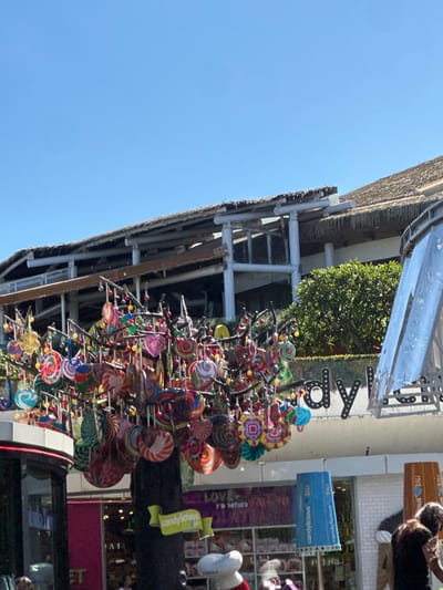 In Kuta, Indonesia, a tree adorned with colorful lollipops stands outside a store. The store's signage is visible, with large letters spelling out "candylicious". The building has a thatched roof and a modern architectural style. A blue cylindrical sign with "candylicious BALI" written on it is prominently displayed. In the background, there are shops and a clear blue sky. A person with long, curly hair is partially visible on the right side of the image.