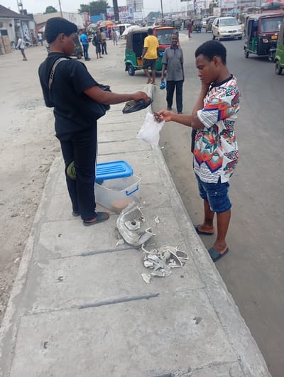 The image portrays an urban street scene in Aba, Nigeria, under daylight conditions. In the immediate foreground, two young males are interacting on a concrete drainage cover adjacent to a paved road. The male on the left, with his back to the camera, is dressed in a black t-shirt, black trousers with white side stripes, and black slippers. A black shoulder bag with a white strap is visible. He extends a black object, resembling footwear, in his left hand towards the male on the right. A blue plastic storage container with a lid is situated on the concrete near his feet. Adjacent to the container, several fractured pieces of a white ceramic or plastic object are scattered on the concrete surface.
The male on the right, facing the first individual, wears a short-sleeved shirt with a multi-colored geometric pattern, ripped blue denim shorts, and black slippers. He extends his right hand forward, appearing to receive a small white plastic bag. His left hand is positioned near his head.
The background reveals a bustling street with active vehicular traffic, including multiple green and yellow tricycles and various cars, such as a white sedan. Several pedestrians are visible walking on the street and sidewalks. Buildings with commercial fronts and overhead utility lines line the street, with billboards discernable in the distance. The sky is overcast.
