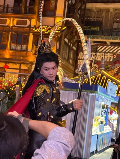 A person in ornate black and gold armor with a red cape stands holding a large, ornate spear. The person has dark hair pulled back and is looking towards the viewer. The background features a building with traditional Chinese architecture, as well as a street scene with various storefronts and neon lights. The location is Yuzhong District, China.