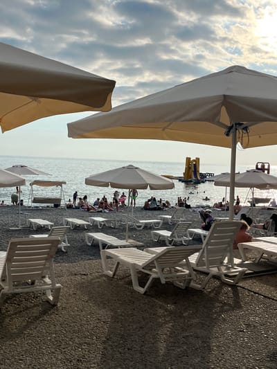 This is a photograph taken on a beach in Sochi, Russia. The scene depicts a lively beach with numerous people enjoying the water and sun.
The main subjects are the beachgoers, scattered across the pebble beach and in the shallow waters of the Black Sea. They are engaged in various recreational activities, including swimming, lounging on beach chairs, and playing. A yellow and blue inflatable water slide is visible in the sea, adding to the playful atmosphere.
Numerous white plastic lounge chairs are set up on the beach, many occupied by people or casting shadows. Several large beige beach umbrellas provide shade, with two prominently in the foreground, partially obscuring the view. The sea is calm, reflecting the bright sky.
The time of day appears to be late afternoon or early evening, as indicated by the quality of light and the long shadows cast by the chairs and umbrellas. The weather seems pleasant, with a partly cloudy sky where sunlight breaks through, creating a somewhat dramatic effect. The pebbly texture of the beach is a characteristic detail of many Black Sea coastlines. The overall mood is one of relaxation and enjoyment.
No visible text is present in the image.