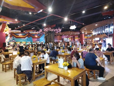 A high-angle shot shows the interior of a bustling public space with tables and seating, identified as Hong Gai Ward, Vietnam. Numerous people are present, seated at wooden tables, engaged in various activities such as eating, talking, and using mobile devices. Each table is decorated with a small vase of yellow flowers. The seating consists of small, square, wooden stools. Overhead, the ceiling features a complex design with various architectural elements, including beams, lighting fixtures, and decorative structures like faux cherry blossom trees. In the background, more people can be seen milling around, suggesting a busy food court or event space. The overall lighting is bright, contributing to the vibrant atmosphere of the place.