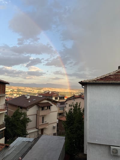 The image depicts a view of Sandanski, Bulgaria, with a rainbow arcing across a cloudy sky. The foreground is dominated by rooftops of residential buildings, characterized by red tile roofs and light-colored facades. Some of these buildings appear to be under construction or in a state of disrepair, with exposed brickwork and unfinished sections visible.
In the midground, more houses and buildings are visible, interspersed with green trees. The buildings are a mix of architectural styles, with balconies and different colored walls.
The background shows a sprawling urban landscape extending towards a range of mountains under a hazy, possibly dusty, sky. The light suggests it is either early morning or late afternoon, with the sun casting a warm glow on the distant landscape. The presence of small white specks scattered throughout the image could indicate falling rain or snow, consistent with the phenomenon of a rainbow. There are no people visible in the image.