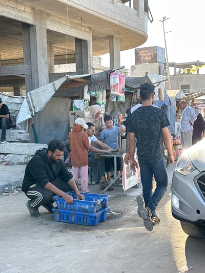 The image depicts a street scene in Gaza, Palestinian Territories, likely during the daytime given the bright sunlight casting shadows. The setting appears to be an informal marketplace or a roadside stall, with makeshift structures and various goods displayed.

Several people are visible, engaged in activities that suggest trade or daily life. In the foreground, a man with a beard, wearing a black hoodie and track pants, is sitting and looking to his left. Next to him, a young person in a light blue t-shirt is also seated. Further back, another man in a dark patterned t-shirt and jeans is standing, his arms crossed. Other individuals are seen in the background, some wearing traditional headscarves.

Objects in the scene include stacked blue plastic crates, bags of what appear to be toiletries or household items, and possibly food items like apples on a cart. There's also a silver car parked on the right side of the image. Construction materials, such as rebar and concrete, are evident in the partially built structures on the left, indicating ongoing development or perhaps damage.

A large billboard or banner with Arabic text is visible in the background, featuring the word "FESTIVAL" and what appears to be "Ajman." The overall atmosphere seems industrious, with people gathered in an outdoor commercial area. The weather appears to be sunny and warm.