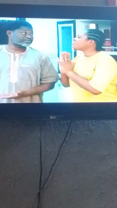 The image shows a still from a television screen depicting two individuals. On the left, a man with dark skin and graying facial hair is wearing a light brown garment with a decorative neckline. He is looking to his right. On the right, a woman with dark skin and braided hair is wearing a yellow shirt. She has her hands clasped together in front of her chest, as if in prayer or supplication, and she is looking to her left. The background appears to be an indoor setting with a doorway visible. The image is framed by the television screen and the lower portion shows the surface of the television stand, which has some cables visible. The specific location in Lokoja, Nigeria, cannot be determined from the visual information presented in the image.