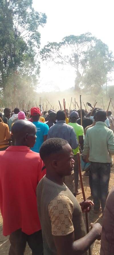 The image shows a crowd of people standing in a clearing in a forest, located in Malava, Kenya, at the coordinates 0.427 latitude and 34.815 longitude. There is a lot of dust in the air, which appears to be kicked up by people who are walking towards the camera. Many of the people are holding wooden staffs. The man in the foreground is wearing a red shirt. The man to his right is wearing a green shirt with a white inscription and a red hat. The man in the center of the image is wearing a green shirt with a brown "Gucci" pattern on the sleeves. The man in the background, to the left of the center, is wearing a gray shirt. The man to the right of the center is wearing a green shirt. Most of the people are looking in the direction of the camera. The image was taken in the daylight with the sun in the background.