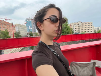 A young woman with dark hair and sunglasses is standing on a bright red pedestrian bridge. She is wearing a dark brown t-shirt and a black crossbody bag with a crocodile-embossed strap. In the background, there are buildings, some under construction with cranes, and trees. The bridge overlooks a canal or river. The image appears to have been taken in Pilsen, Czechia, specifically on the pedestrian bridge connecting the city center to the Lochotín district, known for its modern red design.