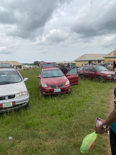 The image depicts an outdoor scene in Durumi, Nigeria, characterized by a cloudy sky and a grassy field with several vehicles. A silver sedan is parked on the left, with its hood open, and a red hatchback is positioned to its right, also with its trunk and driver's side door open. A maroon station wagon is further to the right, with its rear hatch open. Several people are visible near the vehicles, some appearing to be engaged in activities around the cars. In the background, there are buildings with light-colored roofs, suggesting a semi-urban or rural setting. A person in the foreground on the right is holding a green plastic bag and a booklet. The overall impression is one of a gathering or event where vehicles are being utilized for transport or storage.