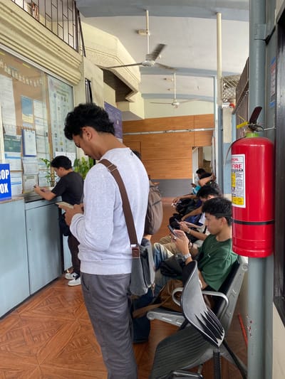 The image depicts a waiting area, possibly at a government office or a service center in Davao City, Philippines. In the foreground, a man wearing a light gray sweatshirt and gray pants stands facing away from the camera, looking at his phone. He has a brown strap across his chest. To his left, behind a counter with various documents displayed, another man in a black t-shirt is seen. A potted plant is also visible on the counter.
A line of people are seated on metallic chairs in the background, all seemingly engaged with their mobile phones. To the right of the seated individuals, a bright red fire extinguisher is mounted on a gray pole. The environment features wooden paneled walls and a tiled floor with a geometric pattern. Ceiling fans are visible on the ceiling, suggesting a warm climate. The overall impression is one of a public space where people are waiting for a service.