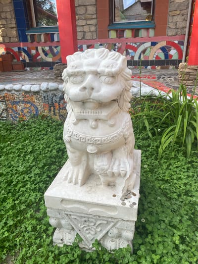 The image shows a white stone lion statue, a common architectural element found outside buildings in Asian cultures, placed in a garden. The lion is seated on a decorative pedestal and is surrounded by lush green clover. Several coins are scattered on the pedestal. In the background, a vibrantly decorated building with colorful mosaic tiles and red architectural elements can be seen, suggesting a location with a unique and eclectic style. The building appears to be a private residence or a culturally significant site. The overall scene suggests a well-maintained outdoor space with an artistic and cultural flair.