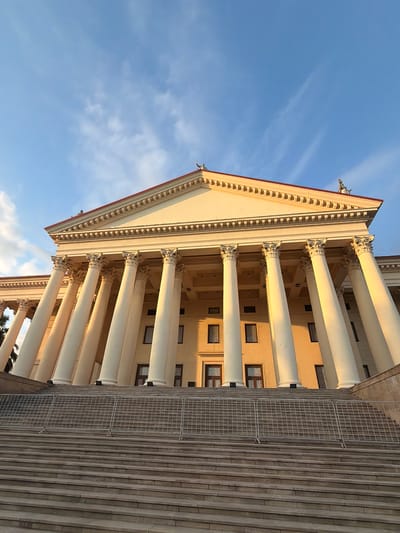 The image shows the facade of a grand, neoclassical building in Sochi, Russia, specifically the State Museum of the History of Religion. The building features a prominent portico with a series of large, fluted Ionic columns supporting a triangular pediment. The facade is painted a pale yellow, and the roofline is accented with a red trim. Two small, dark-colored statues of figures in classical attire are visible atop the pediment. The building is accessed by a wide, stone staircase, which is partially obscured by a temporary metal barrier at the top. The sky above is a clear blue with a few wispy white clouds, and the setting sun casts a warm, golden light on the building's columns and facade.
