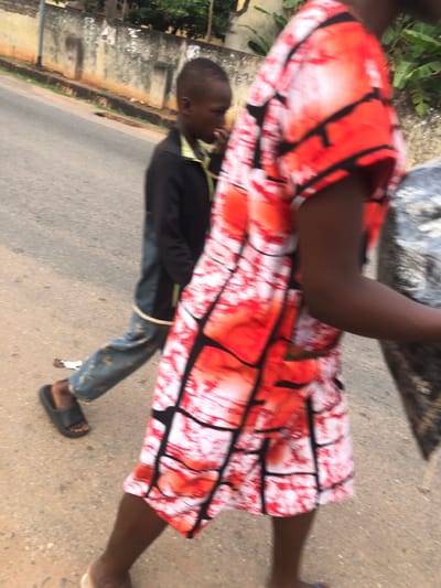 An image shows a child wearing a dark jacket, jeans, and sandals walking on pavement alongside a woman. The woman is wearing a short dress patterned with red, white, and black designs. The image is slightly blurry, and the background shows buildings and greenery. The shot appears to have been taken from an elevated viewpoint.