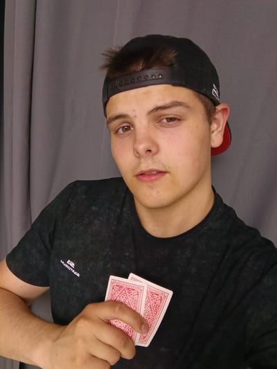 A young man is posing indoors. He has short brown hair and fair skin. He is wearing a black baseball cap with a red brim and a black t-shirt with a small white logo on the left chest. In his left hand, he holds two playing cards with red designs. The background consists of a gray curtain. There is no evidence to confirm the location is Andreyevo, Russia.