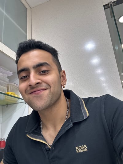 A man smiles at the camera, wearing a black collared shirt with "BOSS HUGO BOSS" embroidered in gold. He has short dark hair and a silver chain necklace. A gold hoop earring is visible in his left ear. The background is a glossy white surface, likely a countertop or wall, with overhead lighting. A glass shelf and some containers are partially visible in the foreground. The image is slightly tilted.