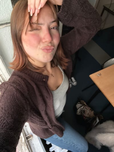 A woman lies on her back wearing jeans, a gray shirt, and a brown knit sweater. Her hair is light brown. She's pouting at the camera. A small cross tattoo is visible on her neck. A desk with a light brown top sits above her. Beside her are a purse, a pair of sneakers, and a fluffy white dog. The scene is angled, as if the picture were taken from above.