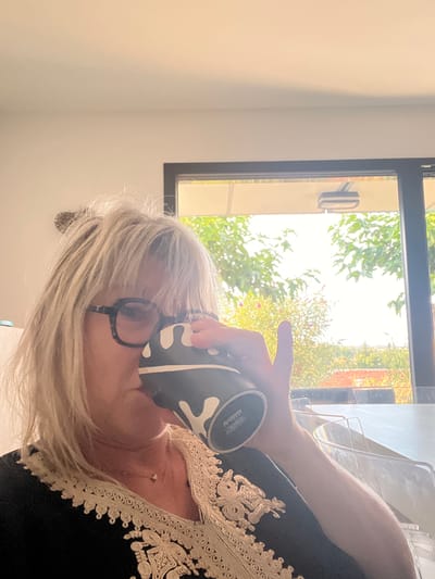 A woman with blonde, messy hair and black-rimmed glasses is drinking from a black and white patterned mug. She is wearing a black garment with ornate gold trim around the neckline. The background features a large window with a view of lush green trees and a glimpse of terracotta rooftops, suggesting a rural or suburban setting. The lighting indicates it is daytime. There are no discernible landmarks placing the image specifically within Chambœuf, France.