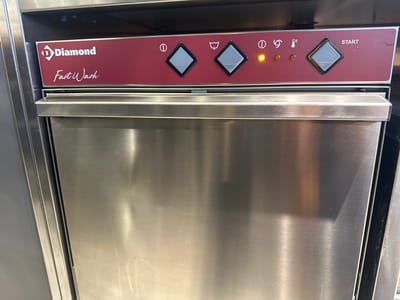 The image shows a close-up view of the control panel of a "Diamond Fast Wash" dishwasher, likely in a commercial setting in Bishkek City, Kyrgyzstan. The panel is a dark red color with white text and symbols.
The subjects are the dishwasher's control panel and the stainless steel appliance itself. The control panel features several buttons and indicator lights. From left to right, there is a circular button with a vertical line inside (likely a power button), followed by a diamond-shaped button. Next is an icon depicting wavy lines, possibly representing wash cycles, followed by another diamond-shaped button. To the right of these, there is a circular button with a vertical line inside, an illuminated amber light, and a thermometer icon with horizontal lines, indicating temperature. Further to the right, there is a large diamond-shaped button and the word "START" in white capital letters. The amber light appears to be on.
The "Diamond" logo is prominently displayed on the left side of the panel, with the words "Fast Wash" written below it in a stylized script. The overall scene is clean and functional, typical of a kitchen appliance. There are no people visible in the image. The lighting appears to be artificial, and there are no indicators of the time of day or weather conditions. The location context is provided as Bishkek City, Kyrgyzstan, but there are no specific visual cues in the image to confirm this. The prominent text includes "Diamond," "Fast Wash," and "START."