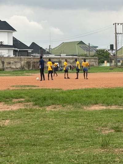 The image depicts an outdoor scene on a cloudy day in Abuja, Nigeria. A group of young men are gathered on a reddish-brown dirt field, appearing to be engaged in a casual game of soccer. Several individuals are wearing yellow and blue athletic shirts and shorts, while one person stands out in a dark blue shirt and black pants. A soccer ball is visible on the ground near the center of the field. In the background, a concrete wall separates the field from a residential area. The structures in the background include modern houses with dark-colored, pitched roofs, some equipped with solar panels, and utility poles with electrical wires. The foreground is dominated by patches of green grass.
