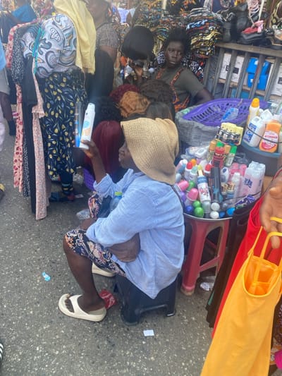 The image captures an outdoor market scene in Accra, Ghana. The foreground features an individual with dark skin, wearing a light blue striped shirt, patterned trousers, white slide sandals, and a light brown woven hat. This person is seated on a black plastic stool, facing left, holding a white spray bottle with a blue label in their right hand. Adjacent to them on the right, a red plastic stool supports a silver tray containing numerous small bottles and containers, alongside larger bottles, one marked "AFTERWASH." Behind this setup, a second individual with dark skin and dark, disheveled hair is seated, wearing a dark top, and holding a short black wig in their left hand. This second person is positioned behind a display of multiple wigs in varying colors, including black, red, and brown. To the immediate left, a clothing rack displays various garments. The ground is dark asphalt with scattered debris. The background is densely packed with market stalls, displaying textiles and various goods, indicating a crowded commercial environment.