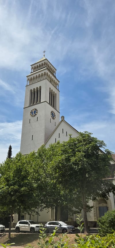 A white church tower with a golden roof stands tall against a blue sky with wispy white clouds. The tower has two visible clock faces with blue backgrounds and golden Roman numerals, one pointing to approximately 10:10 and the other to approximately 10:12. Below the clock faces, the tower features arched windows. Adjacent to the tower is a white building with a sloped roof, also displaying several small, rectangular windows. In the foreground, a dense canopy of green trees partially obscures the view of the church. The ground level shows two parked cars: a white SUV and a silver sedan. The environment appears to be an urban setting, likely within the city of Kehl, Germany, given the architectural style and surrounding vegetation.