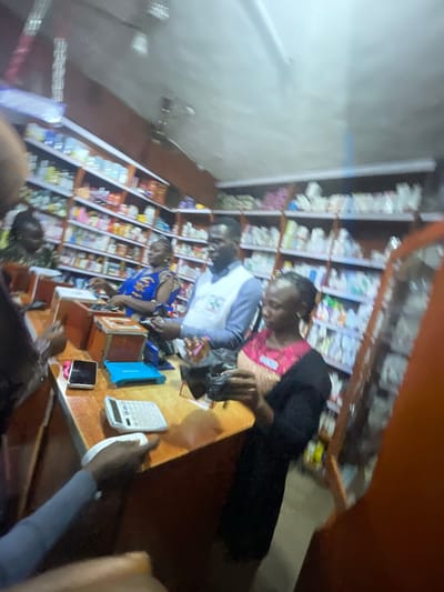 This image depicts an interior scene, likely a small pharmacy or shop, given the shelves stocked with various items, possibly medications. The lighting suggests it could be evening or the interior is dimly lit. Three individuals are visible. In the foreground, a person in a blue and yellow patterned garment is interacting with another person who has on a pink and black outfit. The latter person's head is obscured, but their arm is visible reaching forward. A third person, wearing a blue and white long-sleeved shirt, is partially visible. They appear to be looking down.

On a wooden counter in the foreground, several objects are laid out. These include a white calculator, a dark-colored tablet device, and a smartphone. A black plastic bag is also on the counter. The shelves behind the individuals are filled with numerous small packages and boxes, forming a dense backdrop of products. The overall atmosphere seems to be one of transaction or service within a retail environment. The image is oriented with the counter at the bottom right, and the shelves extending from the left to the right and upward, giving a somewhat tilted perspective. No visible text is legible in the image. The location context is Gura Top, Nigeria.