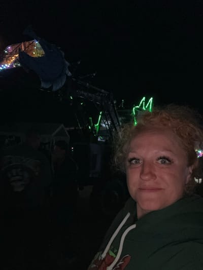 The image depicts a nighttime scene in Большая Кивара, Russia, illuminated by artificial lights. In the foreground, a woman with light-colored, curly hair, light eyes, and prominent eyelashes faces the camera with a neutral to slight smile. She is wearing a dark green hooded sweatshirt with white drawstrings and a visible graphic on the lower left chest. Behind her, a large piece of dark-colored machinery, possibly agricultural equipment, is adorned with bright green LED strip lights, some forming a distinct pattern that resembles an 'M' or crown shape. To the left of the woman and the machinery, multicolored string lights (blue, green, yellow, purple) illuminate a fence or structure, and at least two additional indistinct human figures are partially visible in the darkness. The scene is otherwise dark, with minimal environmental details discernible beyond the illuminated elements.