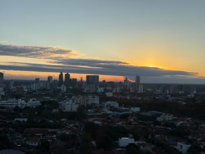 This image captures a panoramic view of the city of Santa Cruz de la Sierra, Bolivia, at sunset. The sky is painted with warm hues of orange, yellow, and blue, with elongated clouds stretching across the horizon. The setting sun is partially obscured by a band of clouds, casting a soft glow over the cityscape. In the foreground, a dense arrangement of residential buildings with red-tiled roofs is visible, interspersed with lush green trees. Further in the background, a cluster of modern high-rise buildings forms the city's skyline, characterized by their geometric shapes and varying heights. The overall scene depicts a tranquil transition from day to night in an urban environment.