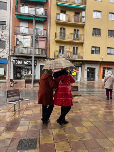 Two women are standing on a tiled sidewalk in the city of Segovia, Spain, near the coordinates 40.939, -4.115. The women are wearing winter coats and are holding a plaid umbrella over their heads. The woman in the red coat is facing the camera. The woman in the brown coat is facing the side. The woman in the red coat is holding the umbrella. The sidewalk is wet, and there are buildings on both sides of the street. The woman in the red coat is wearing black boots. The woman in the brown coat is wearing black boots. The sidewalk is made of large square tiles. There is a bench on the right side of the image. The wall of a building in the background has "Moda Actual" written on it. The woman in the red coat is holding the umbrella with her left hand. There is a large metal drain cover in the lower right corner of the image. The building on the left side of the image is a yellow building. The building has many windows. The building has a brown awning over the top of it. There is a sign in the window on the first floor of the building on the left that says "Romero". There is a red sign in the window on the second floor of the building on the left that says "Rebajas." The woman in the red coat has short dark hair and the woman in the brown coat has longer dark hair. Both women have on what appear to be black gloves. The woman in the brown coat is holding a black purse over her shoulder. The tiled sidewalk is uneven and sloped. The street is wet. The image is taken from a low angle.
