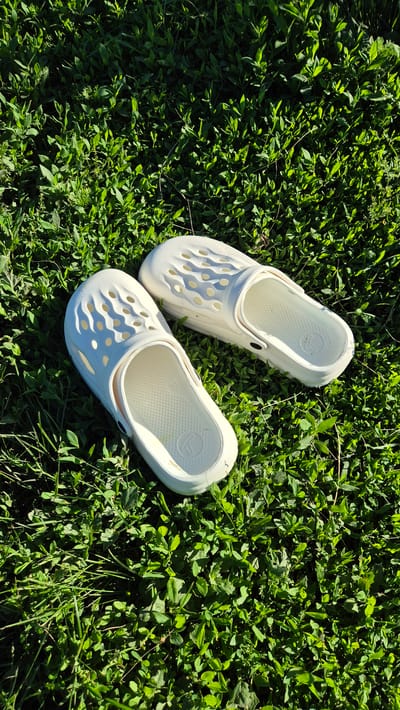 A pair of white clogs are placed on lush green grass. The clogs have a perforated design on the top. The image is taken from a high angle, looking down at the clogs and the surrounding grass. There are no people or any other objects visible in the image. The lighting suggests it is daytime, possibly in the morning or late afternoon, given the shadows cast by the clogs. The environment is a natural outdoor setting. The location is specified as Bulatovo, Russia.