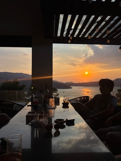 The image captures a sunset scene over Lake Avándaro in Valle de Bravo, Mexico, viewed from an elevated, semi-outdoor terrace. The sun, a bright orange disc, is positioned above the horizon over the lake, casting an intense orange glow across the water and sky. Distant mountains frame the lake on both sides.
The terrace structure includes a dark, vertical architectural pillar on the left side of the frame and a slatted pergola ceiling with multiple strung Edison-style lights visible overhead. A glass railing borders the viewing area.
In the foreground, a dark, reflective rectangular table is set, reflecting the colors of the sunset and clouds. Various objects are arranged on the table: several clear drinking glasses, some appearing used, a red-labeled bottle (likely a soft drink), at least two dark liquor bottles, an ice bucket, a small lit candle in a glass holder, a dark ashtray containing a cigarette butt, a white box (likely a cigarette pack), a pair of red-framed sunglasses, and a wooden tray.
Two individuals are present at the table. On the right, a person with dark, medium-length hair, wearing a t-shirt and shorts, is seated in a dark chair, silhouetted against the sunset, looking towards the lake. Their right leg is crossed over their left. The left arm and a clear glass belonging to a second, unseen person are partially visible in the bottom left corner of the frame. The immediate foreground and interior elements are underexposed, emphasizing the bright sunset in the background.