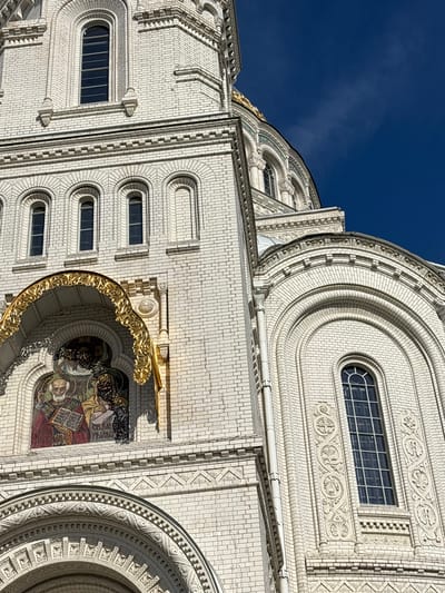 The image shows a close-up, low-angle view of the white brick exterior of a large, ornate building, likely a cathedral, under a bright blue sky with wispy clouds. The architecture features intricate details, including multiple arched windows, decorative brickwork with circular and geometric patterns, and golden decorative elements above a mosaic. One visible mosaic depicts religious figures. The building's facade is brightly illuminated, suggesting it is daytime. The style of the architecture and the presence of religious iconography suggest a location of historical or religious significance, consistent with Kronstadt, Russia.