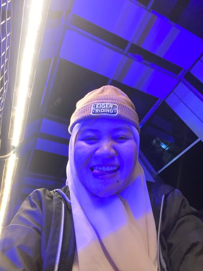 A person is captured in a selfie-style photograph, illuminated by strong blue and white lights. The subject is wearing a light-colored beanie with a patch that reads "EIGER RIDING." They are also wearing a light-colored hoodie or sweatshirt and a dark jacket. The person's face is close to the camera, with their mouth open and tongue visible, suggesting a playful or excited expression. The background is abstract, with panels of blue light and some darker structural elements, possibly indicating an indoor setting with dramatic lighting, like a stage, an entertainment venue, or a studio. There are also bright white lights in the foreground, adding to the artificial lighting environment. The overall impression is one of a candid and vibrant moment, possibly at night given the artificial lighting. The image is oriented sideways.