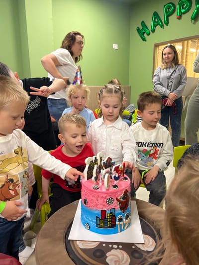 A group of children gather around a Spiderman-themed birthday cake. The cake is half pink and half blue, with a cityscape design on the blue half and Spiderman decorations on top. Several adults are also present, observing the scene. The environment is brightly lit with green walls, and balloon decorations with the word "HAPPY" are on one of the walls. The scene is a birthday celebration, taking place indoors, likely in a party room. It is not possible to confirm the location of Kiryat Bialik, Israel with the given information.