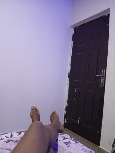 The photo shows legs and feet on a bed with a purple and white patterned sheet. The feet point upwards towards the top of the frame. Behind the legs, there's a pale lavender wall. To the right of the wall is a dark brown door with a silver handle. The door frame is painted white, with some paint splatters visible on the door and wall. The lighting in the room appears to be somewhat dim and cool-toned.
