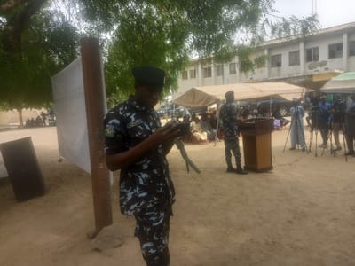 The image shows a scene in Maiduguri, Nigeria, at coordinates 11.869 latitude and 13.125 longitude. A man wearing a camouflage uniform and a green beret is standing on a dirt path, holding a black device in his hands. He is looking down at the device. Behind him, a large white banner hangs from a wooden pole. To the right of the man, a group of people are seated on chairs in front of a wooden lectern under a canopy tent. A person, seemingly a journalist, stands behind the lectern with a microphone in their hand, and several other people are standing behind the lectern, holding cameras and microphones. The background includes a building with windows, tents, and cars. There is a tree on the left side of the image. The scene appears to be an event, potentially a press conference or a public gathering.