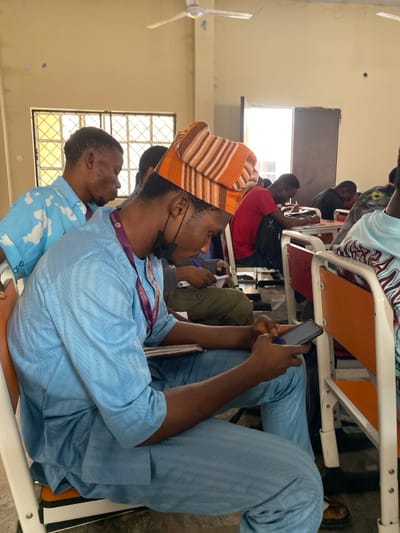 The scene is set inside a classroom or lecture hall in Minna, Nigeria, occupied by multiple individuals, primarily young adult males, seated at desks.

In the foreground, a dark-skinned male is seated in profile, facing right, on an orange chair with a white metal frame. He is dressed in a light blue traditional Nigerian long-sleeved top and matching trousers with a subtle patterned texture. A multi-colored (orange, brown, and beige striped) cap adorns his head. A black face mask is positioned below his chin, and a maroon lanyard with partially visible white text hangs around his neck. He is actively holding a black smartphone with both hands, his gaze fixed on its illuminated screen.

Behind the foreground subject, another dark-skinned male is visible, wearing a light blue short-sleeved shirt with white cloud-like patterns, also looking downwards. Further into the midground, other individuals are seated at similar desk-chair units with white frames and orange or red seating. One individual in a red t-shirt is visible with a black backpack, and others are seen looking down, possibly engaged in writing or similar activities.

The room's walls are painted a faded light yellow or beige, showing signs of wear, including stains and minor surface damage. Two windows with horizontal metal security bars are present on the left side, admitting bright natural light. A white four-bladed ceiling fan is mounted overhead. A partially open doorway on the right side of the frame reveals a brightly lit area beyond. The floor is a dark, unadorned surface.