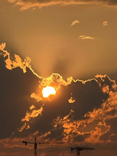 The image captures a dramatic sunset over Minsk, Belarus. The sun, a brilliant orb of white and yellow light, is partially obscured by dark, billowing clouds that are edged with fiery orange and gold. Rays of sunlight pierce through the clouds, creating a striking visual effect. In the foreground, the silhouette of a construction crane is visible against the colorful sky, suggesting ongoing development in the city. The overall atmosphere is one of awe-inspiring natural beauty contrasted with the industrial landscape.
