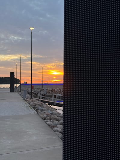 An outdoor, full-shot image captures a waterfront promenade during sunset. The sky is filled with hues of orange and yellow, indicative of a setting sun, with wispy clouds scattered across a pale blue backdrop. In the foreground, a concrete pathway runs along the left side of the image, bordered by a low wall of rounded grey stones and a dark, textured vertical structure on the far right.

Along the pathway, several lampposts stand tall, their lights illuminated, casting a soft glow. In the middle ground, a few figures, appearing as silhouettes, are visible near the waterfront, seemingly engaged in conversation. The water itself is calm, reflecting the colors of the sky. Beyond the immediate promenade, wooden structures suggestive of a pier or docks are present, with some boats or ships visible on the horizon. The overall atmosphere is serene and picturesque, typical of a coastal urban setting at dusk.

This scene is likely located in the city of Tallinn, Estonia, near the harbor area or a coastal promenade. The architectural elements like the lampposts and the dark, textured structure could be part of modern urban development along the waterfront. The presence of a promenade, waterfront access, and potential boat traffic aligns with the coastal geography of Tallinn.