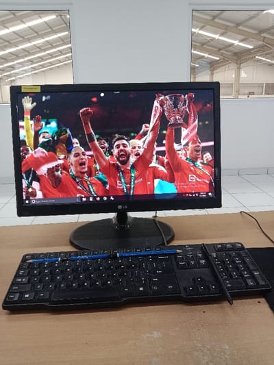 The image shows a computer monitor, keyboard, and number pad on a desk. The monitor is displaying an image of soccer players in red jerseys celebrating a victory with a trophy. Two blue pencils are laying horizontally on the keyboard, with the back of the number pad resting on the pencil closest to the viewer. The desk is made of a brown wood-like material. The image appears to be taken in an office in Kecamatan Ngaliyan, Indonesia (latitude: -7.01, longitude: 110.364). The background shows an office or a warehouse with white walls and a large window, suggesting a bright and well-lit environment.
