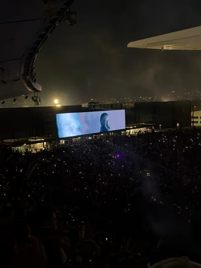 The image shows a nighttime concert scene in Başakşehir, Turkey. A large screen displays an image of a head and shoulders on a cloudy background. A dense crowd of people with their cell phones held up fills the space in front of the stage. The concert appears to be taking place outdoors, with surrounding city lights visible in the distance. A stadium structure is visible on the left side of the frame.