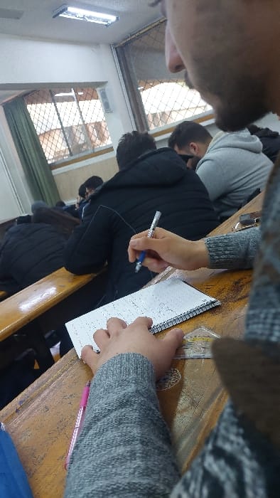 A classroom scene shows students in a row, seated at wooden desks. One student is writing in a notebook with a blue pen, while another holds a pink pen. The perspective is from above, with the back of the head of another student visible. Large windows with wire mesh cover parts of the scene.