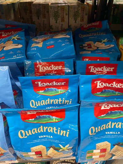 The image shows a retail display of "Loacker Quadratini" vanilla-flavored wafer cubes in blue packaging. The products are stacked on shelves in a grocery store environment. The packaging features the Loacker logo in red, a scenic mountain landscape, and images of the wafer cubes and vanilla pods. Some packages also have text indicating "No. 1 in Italy." The background is blurred, showing other products on shelves, including tea boxes. The scene appears to be an indoor retail setting. There is no specific information in the image to pinpoint the location to Davao City, Philippines; however, it is a common grocery store display.
