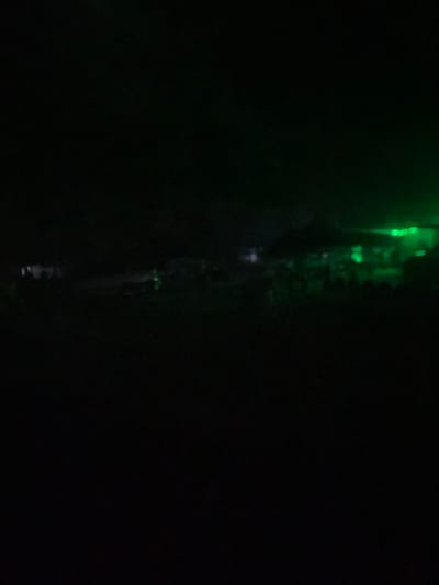 The image depicts a dimly lit scene, possibly an outdoor event or gathering, in Howayi, Nigeria. The majority of the image is in shadow, with the exception of some areas illuminated by green lighting. These lights highlight indistinct shapes that could be structures or a crowd of people. The green light emanates from the right side of the image, casting a glow over the middle ground. The overall impression is one of low visibility and an event taking place at night.