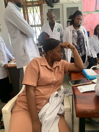 In Dar es Salaam, Tanzania, a group of individuals, some wearing white lab coats indicative of a medical or scientific setting, are gathered. In the foreground, a woman with dark braided hair sits in a white chair, wearing a peach-colored textured outfit. She is looking to her left with her right hand resting on her forehead. On a wooden table in front of her are a mobile phone, a box labeled "WOODEN TONGUE DEPRESSOR," and some papers. Behind her, several other people in lab coats are standing, suggesting a professional or educational environment, possibly a clinic or hospital. The background includes a window with a patterned grill and some filing cabinets or storage units.