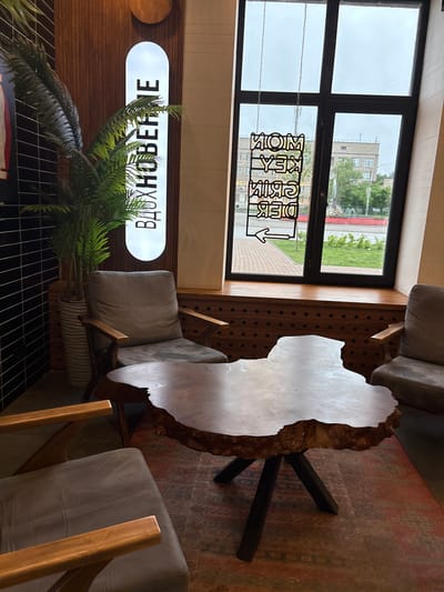 The image displays an interior view of a café or lounge area in Perm, Russia, characterized by a rustic-modern aesthetic. In the foreground, a large, irregularly shaped wooden coffee table with a live edge and dark, angled metal legs rests on a patterned rug featuring red, green, and brown hues. Two armchairs flank the table; the one on the left is partially visible, while the one on the right is almost fully shown, both upholstered in a grey fabric with wooden armrests and frames.
To the left, a tall, striped grey planter containing a large artificial palm plant stands against a dark tiled wall. Behind the planter, a vertical, illuminated white sign with the Cyrillic word "ВДОХНОВЕНИЕ" (Inspiration) in black capital letters is mounted on a brown slatted wooden wall panel.
The right side of the image is dominated by a large window divided into multiple panes by a dark frame, looking out onto an urban street. A black metal sign, suspended by chains from the top of the window, displays mirrored text that appears to read "MOM KEY SKIM DEK" with a left-pointing arrow. Outside the window, an overcast sky is visible above buildings, including a multi-story structure in the distance, and green trees and grass, alongside a street with a fountain. A wide wooden window sill or bench runs along the bottom of the window, beneath which is a wooden panel featuring a pattern of dark circular holes. No people are present in the scene.