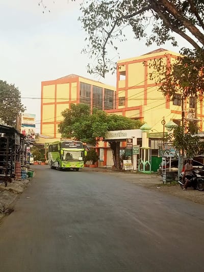The image shows a scene in Pati, Indonesia, located at coordinates -6.754 latitude and 111.039 longitude. A large green and white bus, with "Waisna" written on the side, is driving down a paved road. The bus is in the center of the image, driving away from the viewer. The bus is in the process of passing a smaller white van that is parked on the right side of the road. The road has a gray, asphalt surface and is empty except for the two vehicles. On the right side of the road is a small building with a green roof and a sign above the door that reads "smartfren." In front of the building is a small, open-air market with various vendors selling their wares. On the left side of the road, there is a small wooden building with a thatched roof and a group of people sitting on the steps. The background is a large, three-story building with a yellow facade and a red roof. The building is partially obscured by a large tree with green leaves. The scene is bright and sunny, with the sun shining through the trees. The sky is a clear blue.  The scene appears to be a typical street scene in a small Indonesian town.