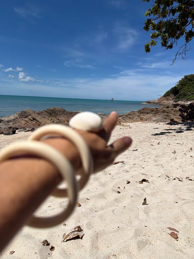 This is a close-up, low-angle, vertical shot of a person's arm and hand at a beach in Baan Plai Laem, Thailand. The person is holding up a seashell and making a peace sign with their fingers. The arm is adorned with a light-colored bangle or bracelet.
The background features a sandy beach with scattered dry leaves and debris. To the left, there are rocky outcrops leading to the ocean. The sea is a calm, turquoise color, and the sky is a clear blue with wispy white clouds. The sun appears to be high in the sky, suggesting it is midday. The scene evokes a sense of relaxation and enjoyment of a tropical environment. No visible text is present in the image.