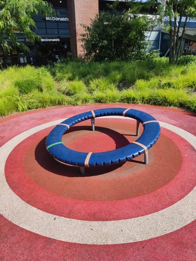 The image depicts an outdoor scene in Rijswijk, Netherlands, featuring a circular merry-go-round as the central object. The merry-go-round is blue and circular, with beige straps holding it together and a green strap also visible. It is supported by metal legs and sits on a red and white circular playground surface. In the background, there is a McDonald's restaurant with its signage visible, surrounded by lush green grass and trees. The architecture of the McDonald's is modern, with large windows and a brick facade. The weather appears to be sunny. No people are clearly visible in the immediate vicinity of the playground equipment, though there are figures inside the McDonald's.