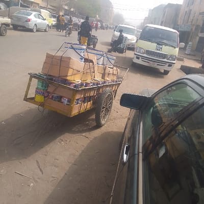 The scene depicts a bustling urban street in Sabon Gari East, Nigeria, under daylight conditions. In the immediate foreground, to the right, a portion of a dark-colored sedan is visible, showing its passenger-side front door, window, and side mirror. Adjacent to it, slightly to the left and centered, a two-wheeled yellow-painted wooden handcart is heavily laden with cargo. The cart's lower section contains various paint cans, identifiable by labels such as "WATER" and "CLASSIC," some appearing yellow and others blue/white. Above these, numerous brown cardboard boxes are stacked, with some visible labels including "MORDEN" and "CLASSIC." The top layer of the cart holds several large white plastic buckets, secured by ropes or straps, along with additional cardboard boxes. The handcart features spoked wheels and casts a shadow to its right.

The road surface in the foreground is unpaved, dusty, and reddish-brown, displaying tire tracks and ground disturbances. Further into the midground, the main thoroughfare is visible, appearing to be a two-lane road with a mix of paved and unpaved sections, indicating wear. Various vehicles populate the road: a silver sedan is visible on the far left, followed by other sedans and vans. Prominently in the mid-right is a light-yellow and white commercial van with a visible Nigerian license plate, facing towards the right side of the frame. Another white sedan is positioned to its right.

Multiple individuals are present on and alongside the road. A person wearing a dark shirt and yellow trousers is seen further down the road, near a smaller cart, suggesting movement or transport of goods. Other pedestrians and at least one individual on a motorcycle or tricycle are visible in the midground. In the background, low-rise buildings with light-colored facades line the street, indicating commercial or residential structures. The sky is hazy and bright, suggesting a sunny day with potentially dust-filled air.